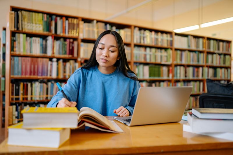 Student writing notes with a laptop and open book in a library