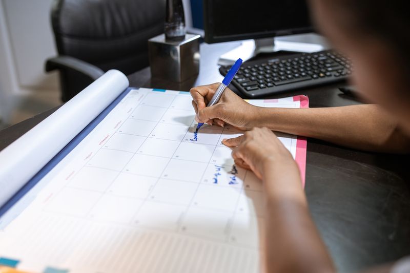Hands writing on a planning calendar while setting up a college application spreadsheet