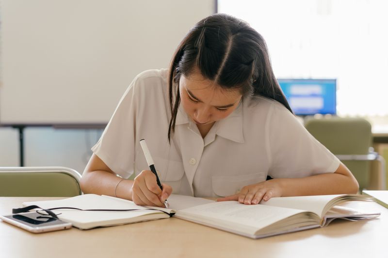 Student writing in a notebook working on a research paper draft